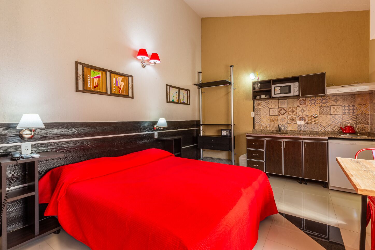 Habitación de hotel con cama, mesita de noche, lámpara, decoración en la pared y kitchenette.
