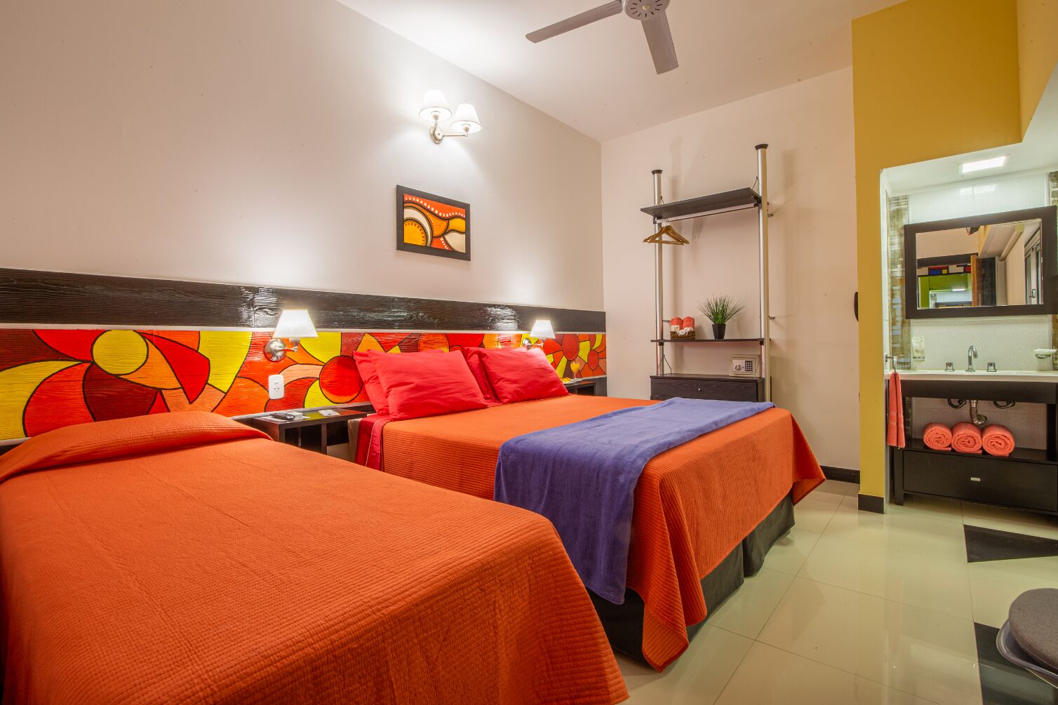 Habitación de hotel con dos camas, ventilador de techo, televisor montado en la pared, mesita de noche con lámpara y baño con lavamanos y espejo.