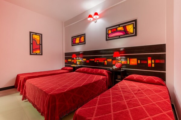 Una habitación de hotel con tres camas, cada una con una colcha roja, un cabecero de madera con un diseño geométrico y dos lámparas fijadas a la pared.