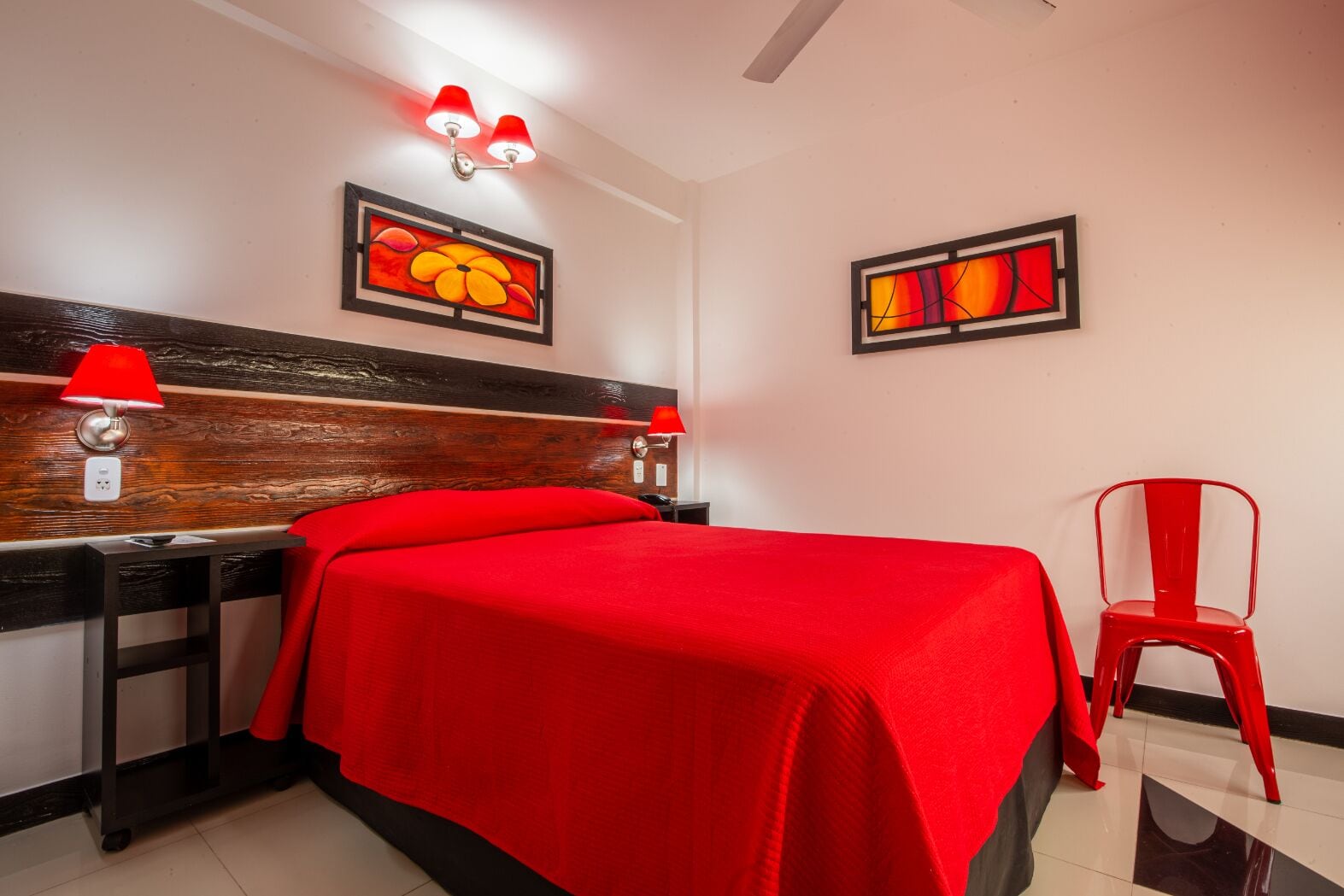 A hotel room with a wooden headboard, a red bedspread, a red chair, and two wall-mounted lamps.