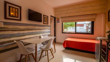 A small, well-lit room with a dining table, two chairs, a bed with a red cover, a TV mounted on the wall, and a window with a view of the outside.