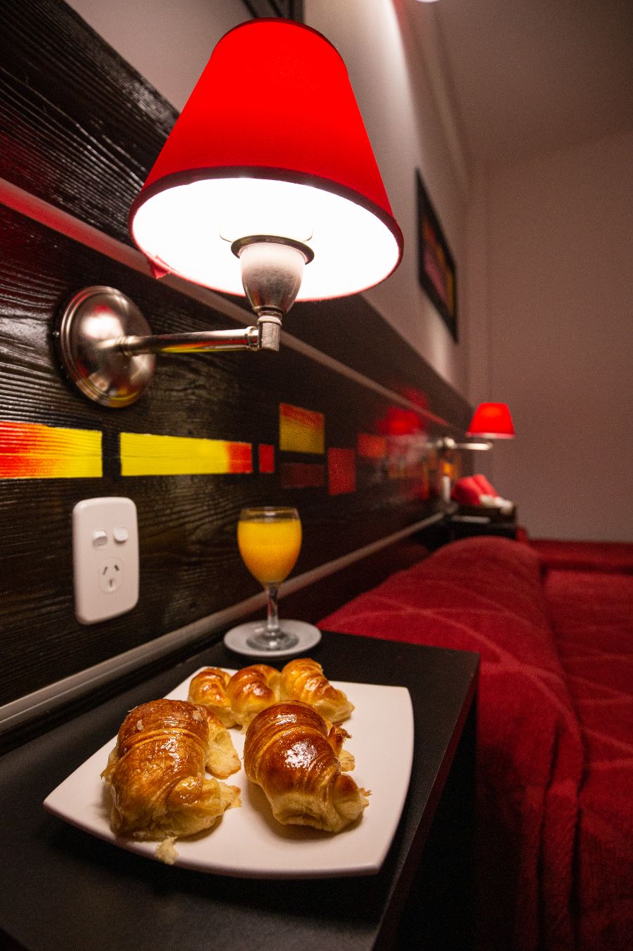 Ein Hotelzimmer mit Nachttisch, Lampe, einem Teller Croissants, einem Glas Orangensaft und einer roten Decke.