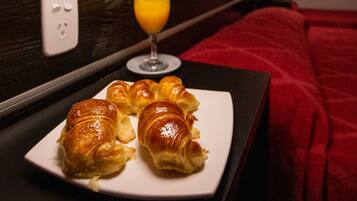 Ein Hotelzimmer mit Nachttisch, Lampe, einem Teller Croissants, einem Glas Orangensaft und einer roten Decke.