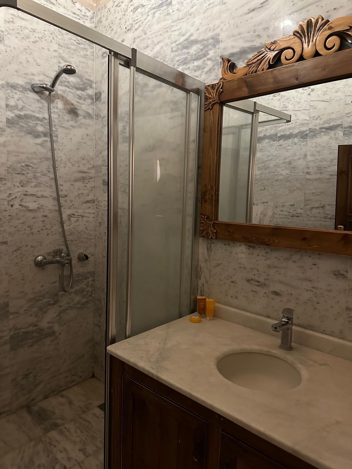 Un baño con cabina de ducha de vidrio, pared con azulejos de mármol, tocador de madera con lavamanos y espejo con marco de madera.