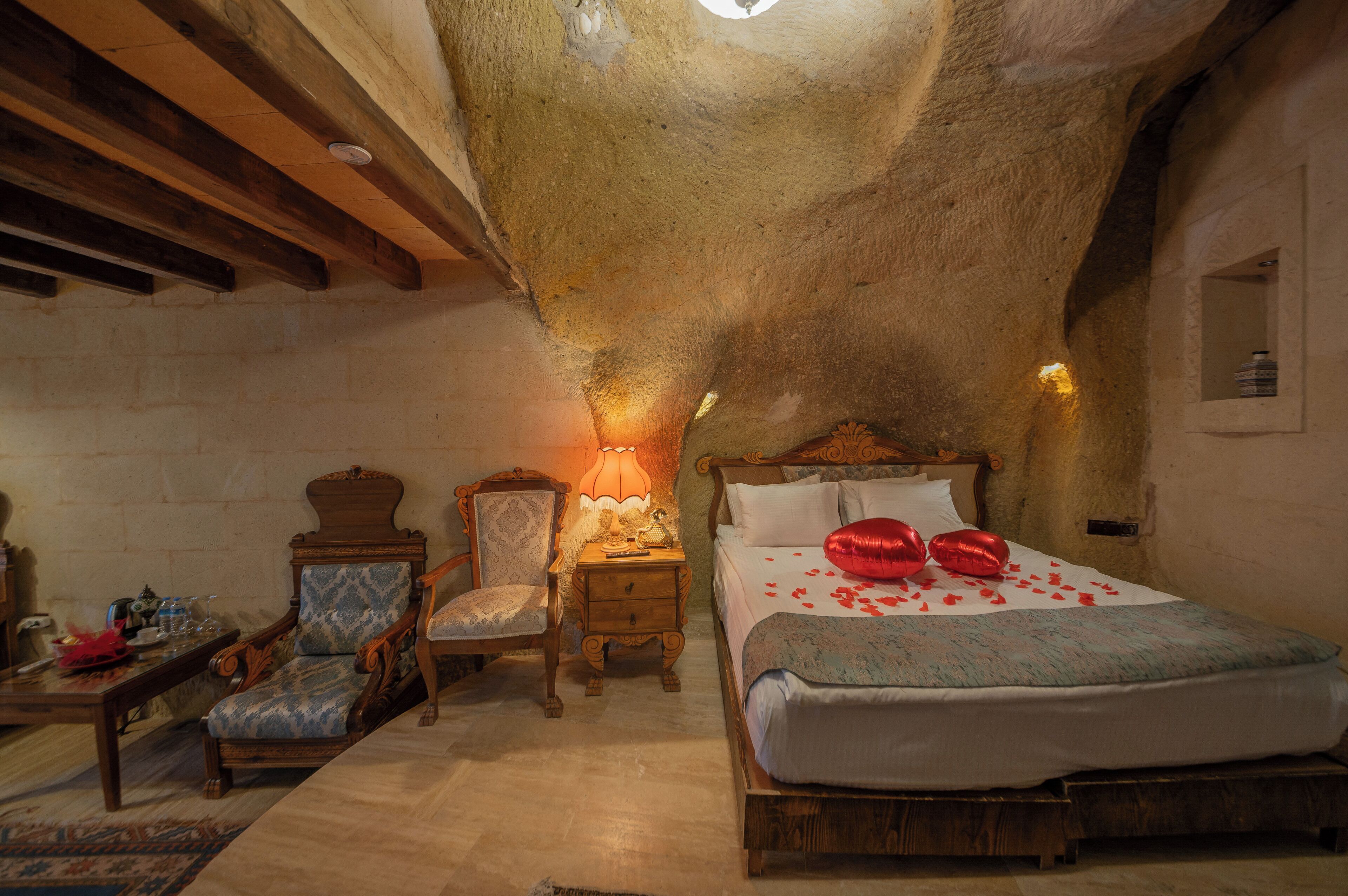 A cozy bedroom with a stone ceiling, a wooden bed, a nightstand with a lamp, and a chair.
