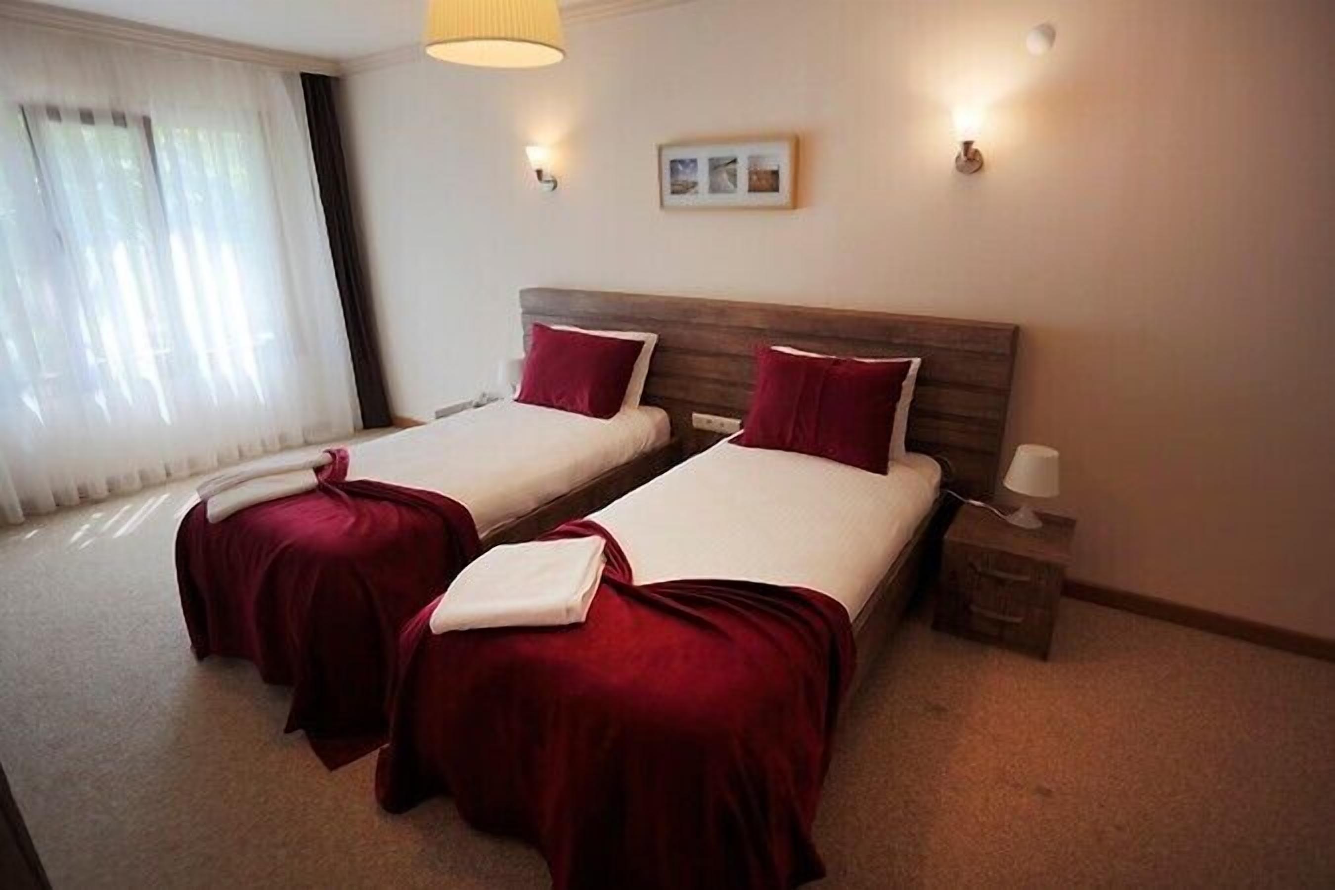 Camera d'albergo con due letti, ognuno con una testiera in legno e biancheria rossa. Sono presenti abat-jour e un piccolo comodino con una lampada.