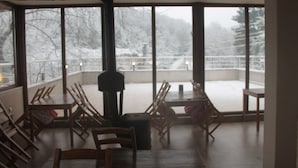 Ein Zimmer mit großem Fenster und Blick auf eine verschneite Landschaft, einem Kamin sowie mehreren Holzsesseln und -tischen.