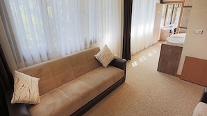 Ein modernes Wohnzimmer mit einem beigefarbenen Sofa, einer kleinen Küchenzeile und großen Fenstern mit durchsichtigen Vorhängen.