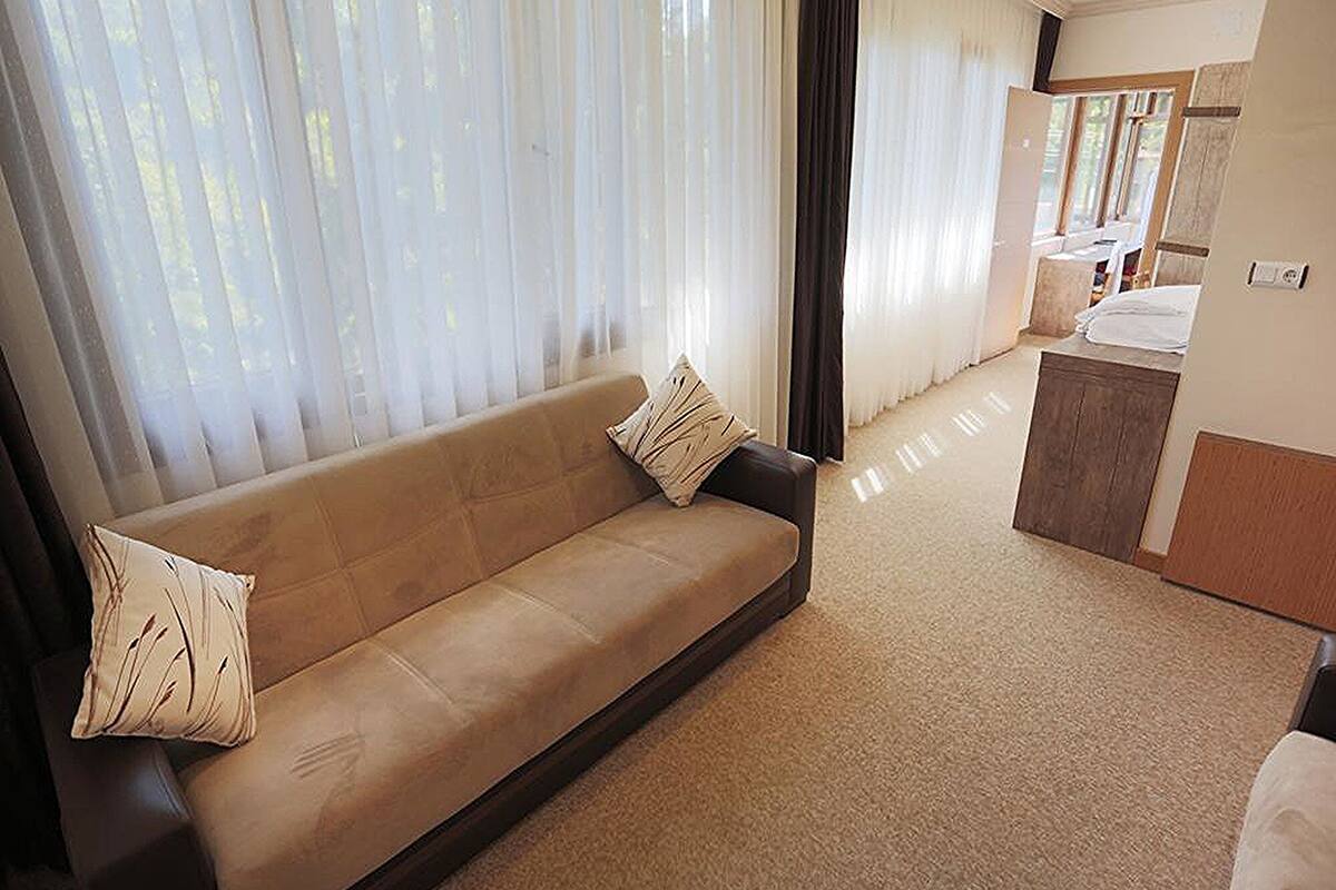 A modern living room with a beige sofa, a small kitchen area, and large windows with sheer curtains.