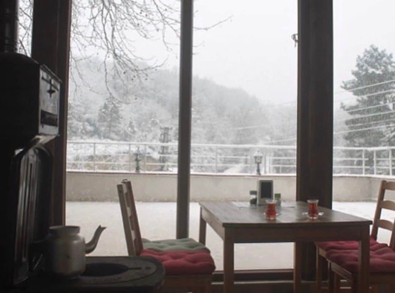 Habitación con vistas a un paisaje nevado a través de amplios ventanales. Se aprecia una mesa con sillas y una pequeña lámpara de mesa.