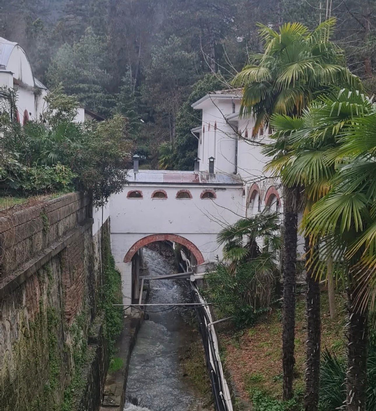 Un étroit canal d’eau passe sous un pont, avec un mur de pierre d’un côté et un bâtiment à toit rouge de l’autre.