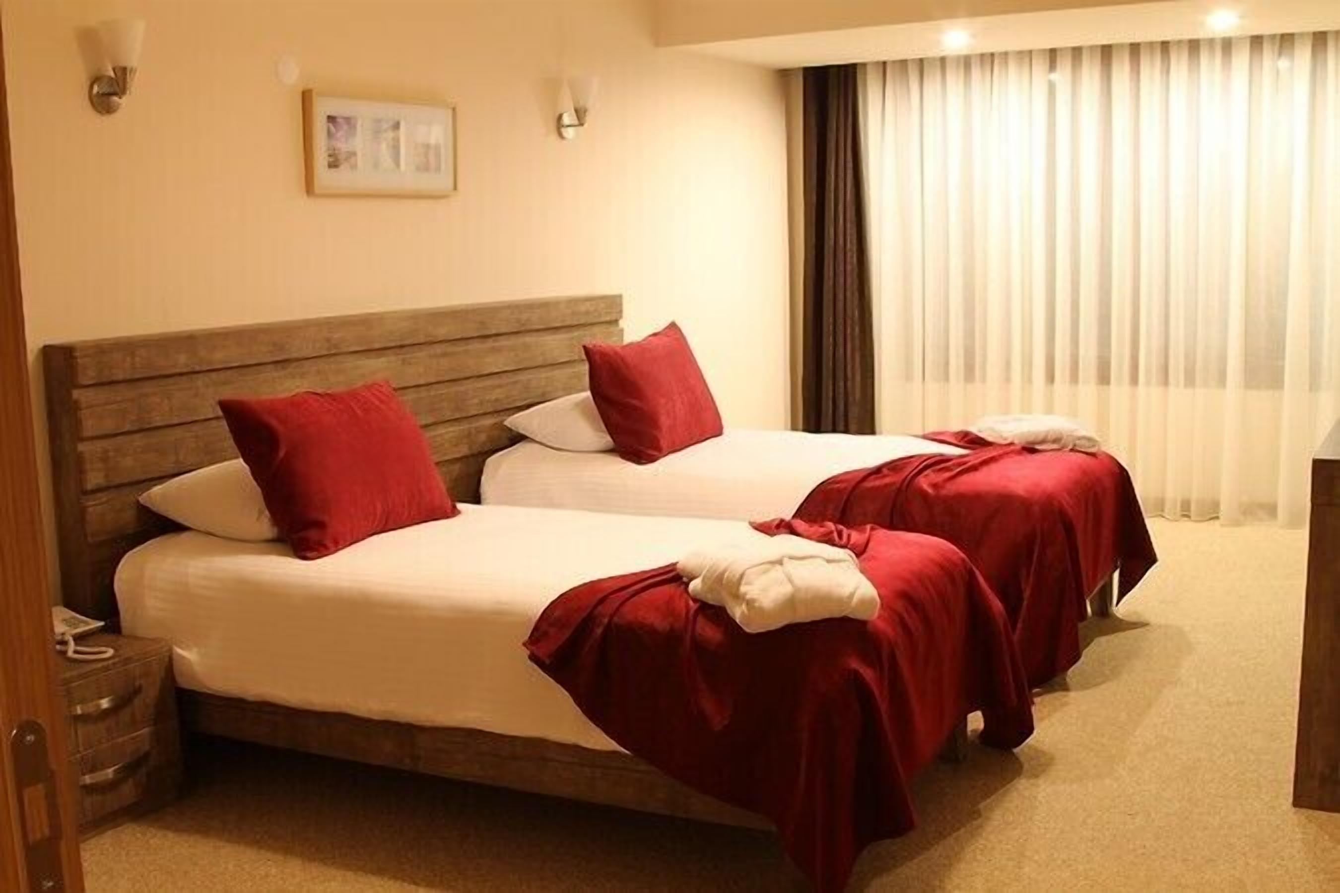 Habitación de hotel con dos camas, cada una con ropa de cama roja y almohadas blancas. Hay apliques en la pared y un cuadro enmarcado.