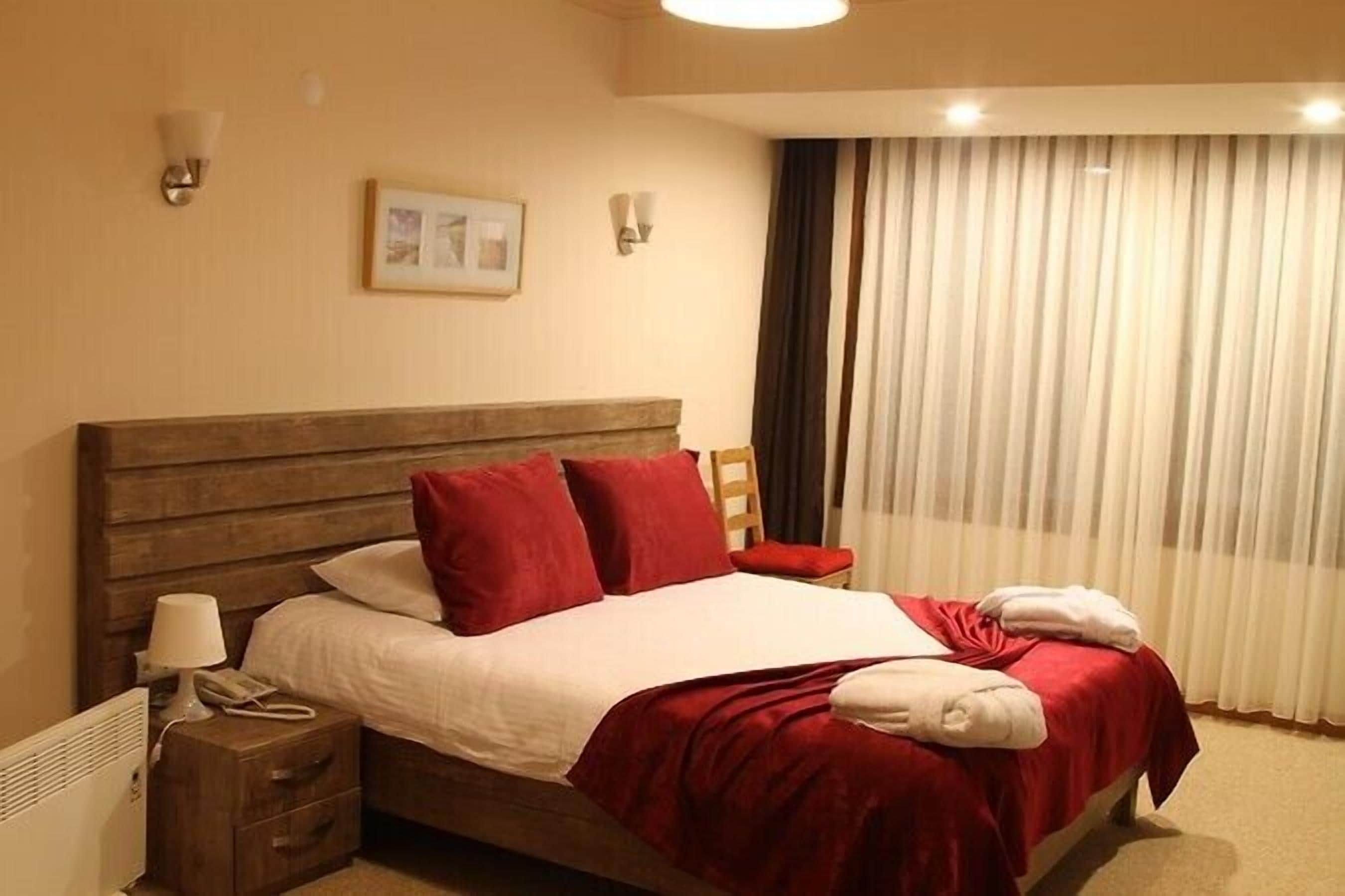 A hotel room with a wooden bed, red and white bedding, a bedside lamp, a nightstand, and a window with curtains.