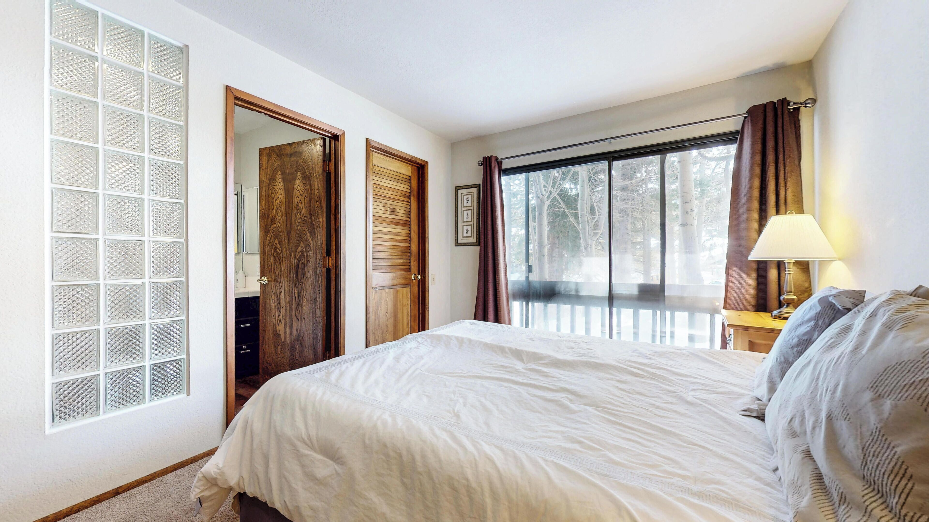 A bedroom with a large bed, a window with curtains, a wooden door, and a nightstand with a lamp.