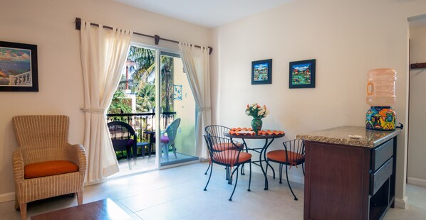 A room with a wicker chair, a dining table with chairs, a water dispenser, and a view through a glass door to an outdoor area with chairs and a table.