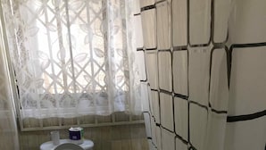 A small bathroom with a toilet, a window with a white lace curtain, and a shower area with a black and white patterned curtain. - The Whitestone Inn (Adentan)