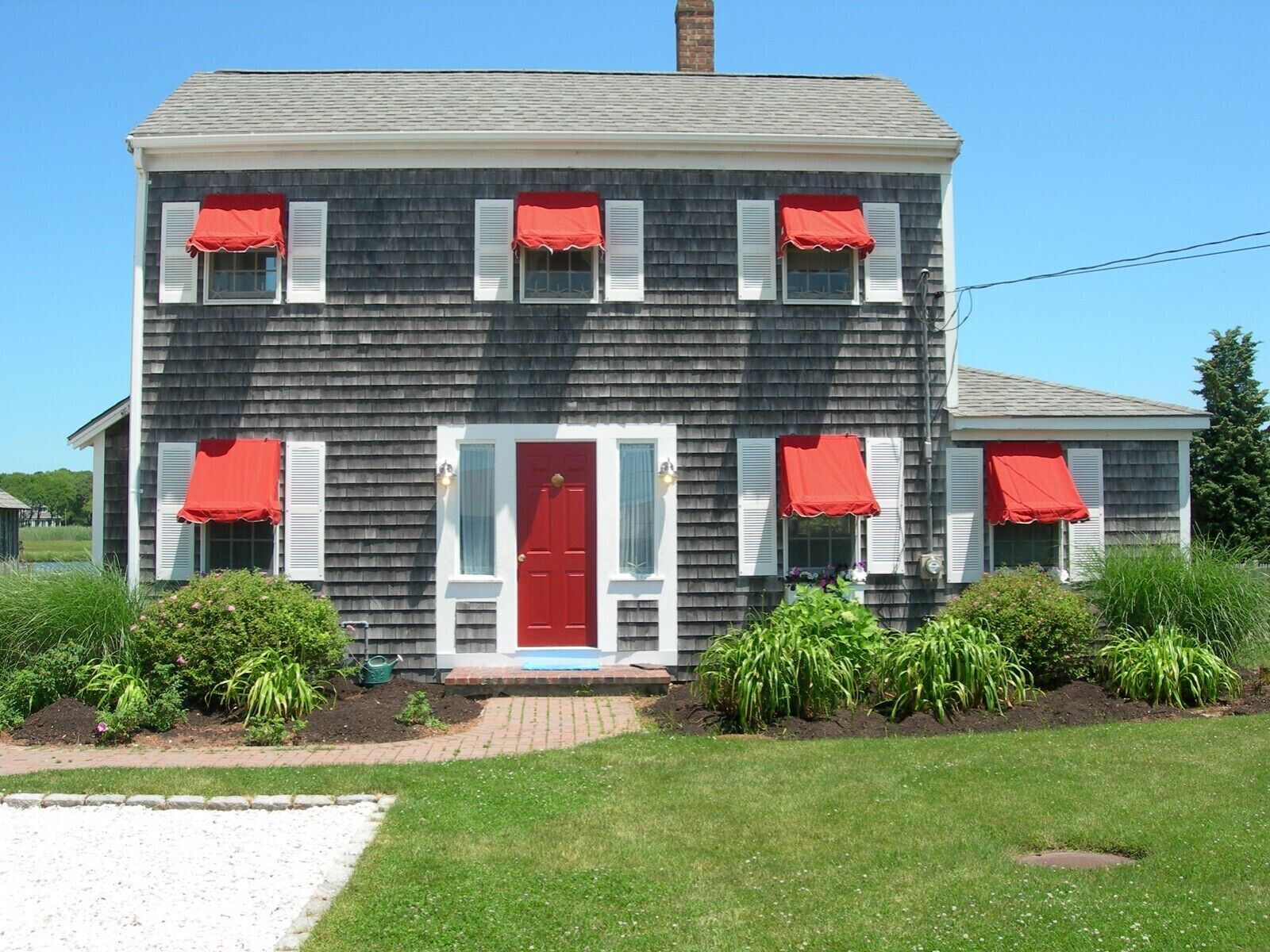 A two-story house with a red door and white shutters, surrounded by greenery and a gravel path.