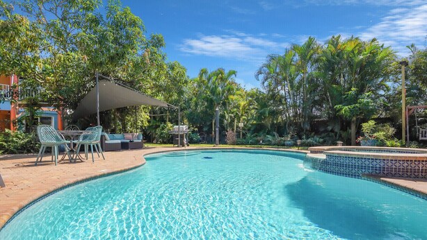 Uma piscina cristalina com borda azulejada, cercada por vegetação e uma área coberta com cadeiras e uma mesa.