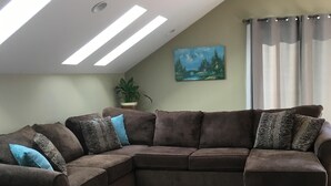 A L-shaped sectional sofa with multiple cushions, a potted plant, and a landscape painting on the wall. - Carlsborg Montian  View Apartment   (Sequim)