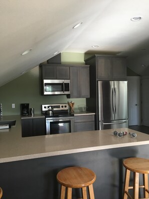 A modern kitchen with a central island, stainless steel appliances, and wooden stools. - Carlsborg Montian View Apartment (Sequim)