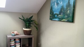 A corner of a room with a wooden floor, a potted plant, a shelf with decorative items, and a painting on the wall. - Carlsborg Montian  View Apartment   (Sequim)