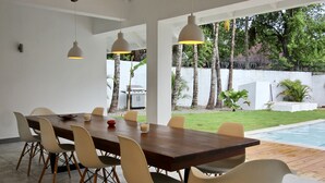 A modern dining area with a wooden table and chairs, pendant lighting, and a view of a swimming pool and greenery. - Casa Panama Modern 5 bedroom villa 250m beach-  (Las Terrenas)