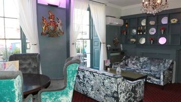 Quarto com sofá de padrão azul e branco, poltrona verde-água e uma pequena mesa redonda. Há um candelabro sobre o sofá e um emblema decorativo na parede.