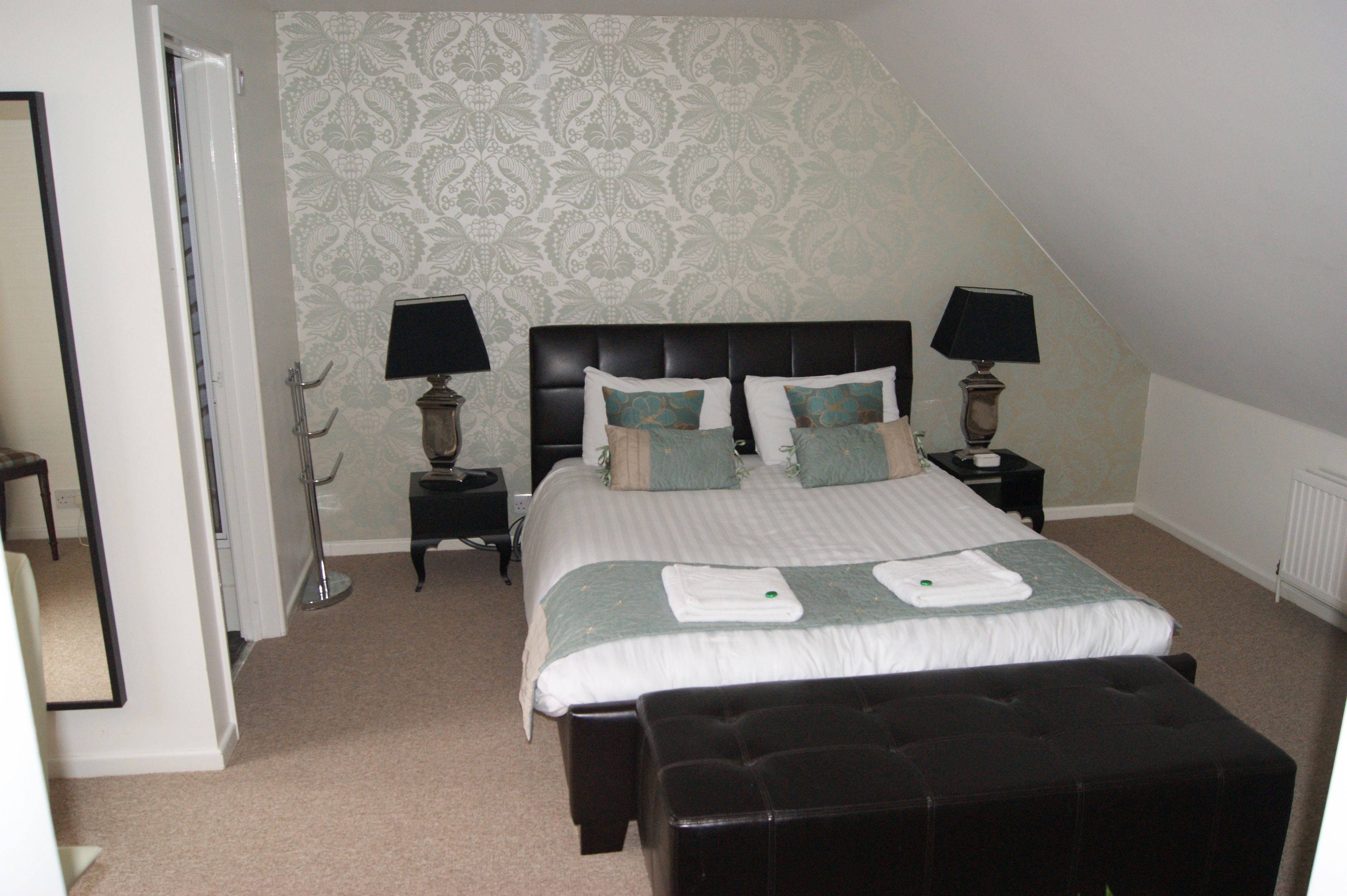 A bedroom with a large bed, two bedside tables with lamps, a bench at the foot of the bed, and a patterned wallpaper on the walls.
