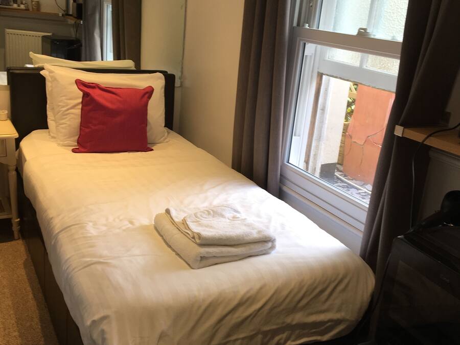 Una cama bien hecha con almohadas blancas y rojas, una toalla blanca doblada y un televisor sobre un soporte a la izquierda.