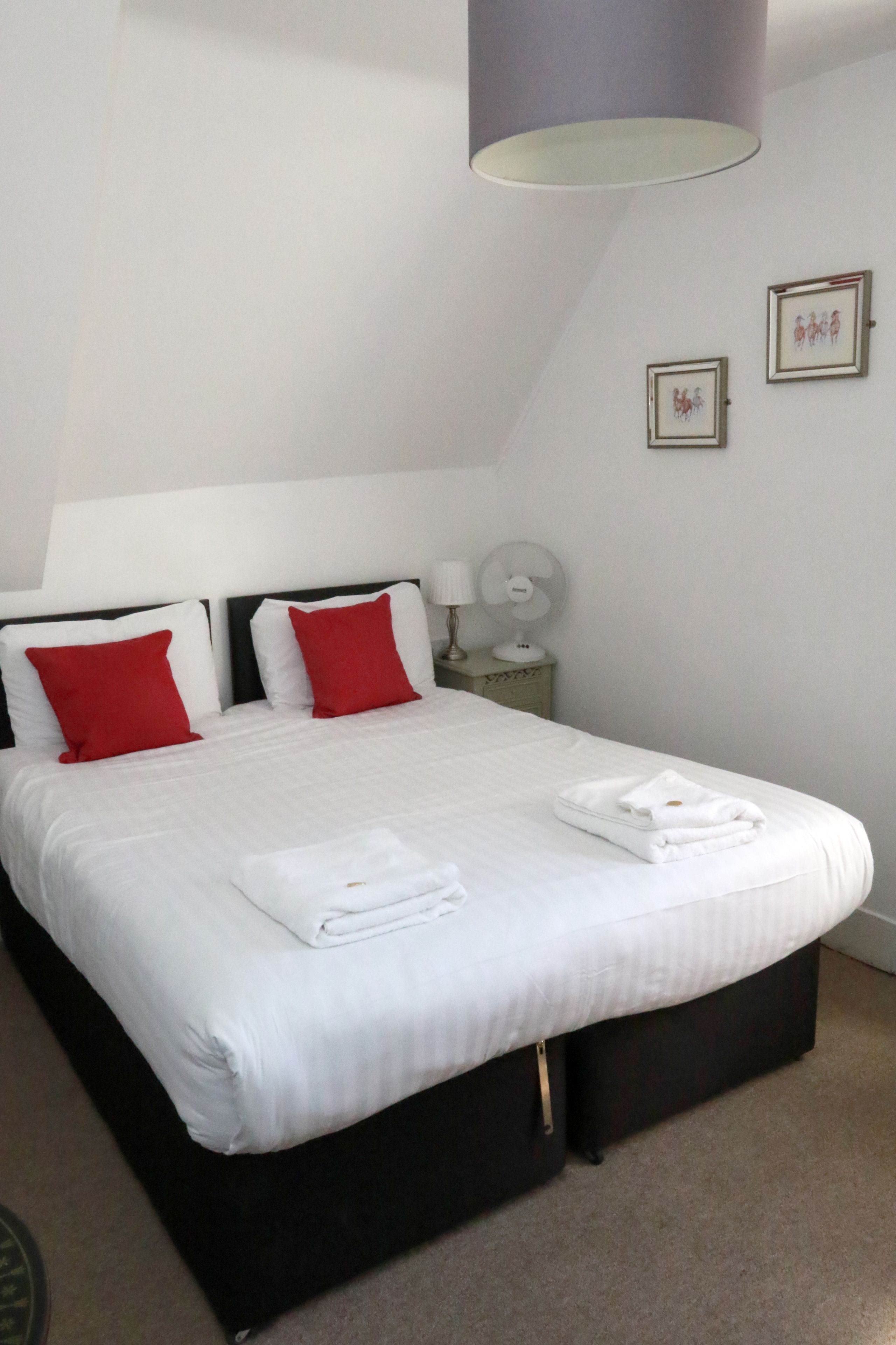 A neatly made bed with white and red pillows, a bedside table with a lamp, and a wall clock.