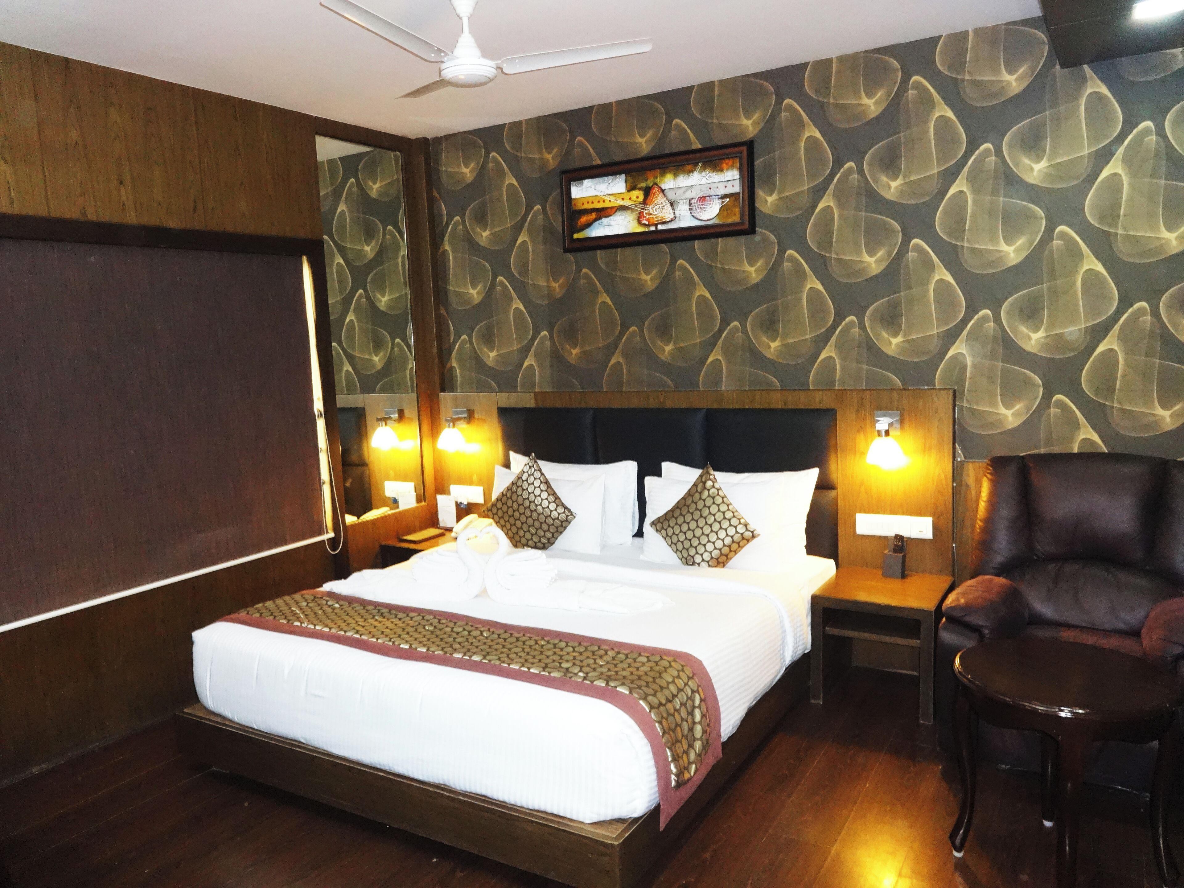 A hotel room with a large bed, wooden headboard, two bedside tables with lamps, a leather armchair, and a television mounted on the wall.