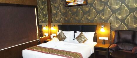 A hotel room with a large bed, wooden headboard, two bedside tables with lamps, a leather armchair, and a television mounted on the wall.