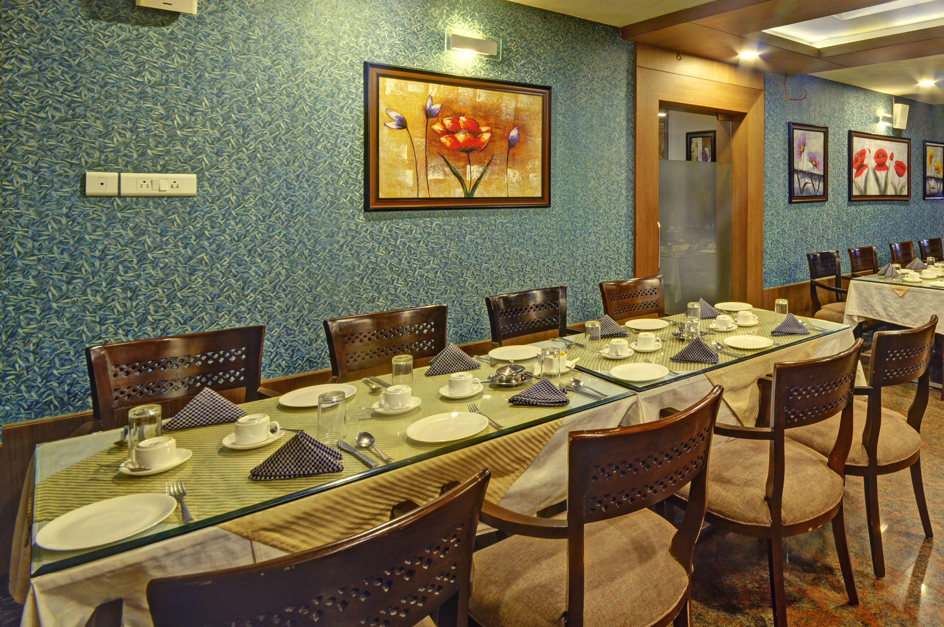 A dining area with a long table set for multiple guests, featuring chairs with intricate back designs, a patterned wall, and framed artwork above the table.