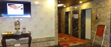 Una hall d'albergo con pavimento in marmo, una reception, un televisore, ascensori e la segnaletica per il check-out.