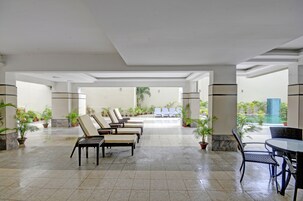 A spacious indoor area with multiple lounge chairs and tables, surrounded by potted plants and a view of an outdoor pool area.