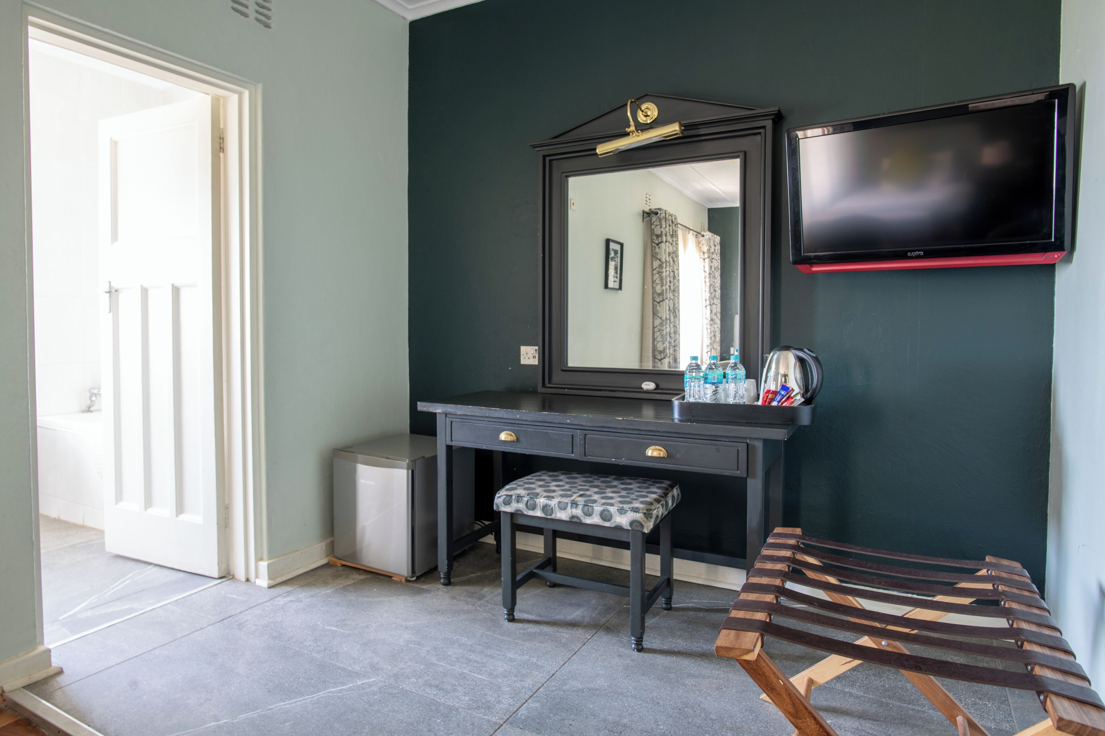 Een kamer met een donkergroene muur, een zwart nagelmeubilair met spiegel, een klein bankje en een televisie boven het nagelmeubilair.
