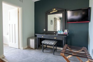 Una habitación con pared verde oscuro, un tocador negro con espejo, un pequeño banco y un televisor montado sobre el tocador.