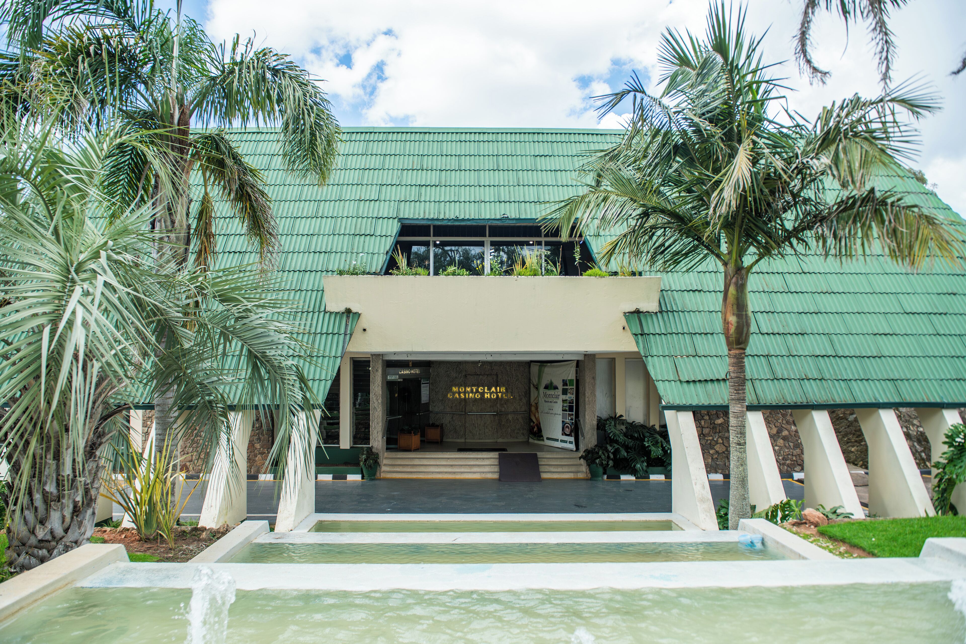 A hotel entrance with a green roof, palm trees, and a water feature in front.