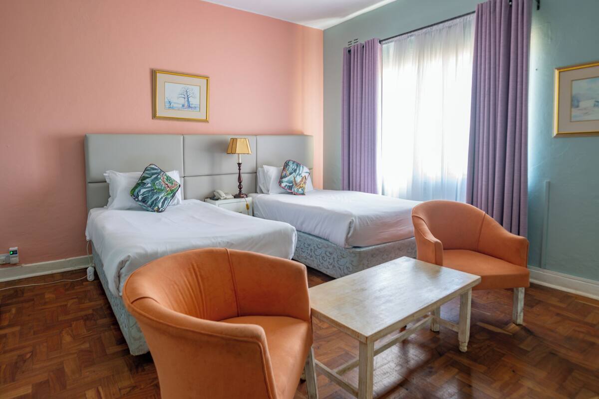 A hotel room with two beds, a nightstand with a lamp, a table, and two orange chairs.