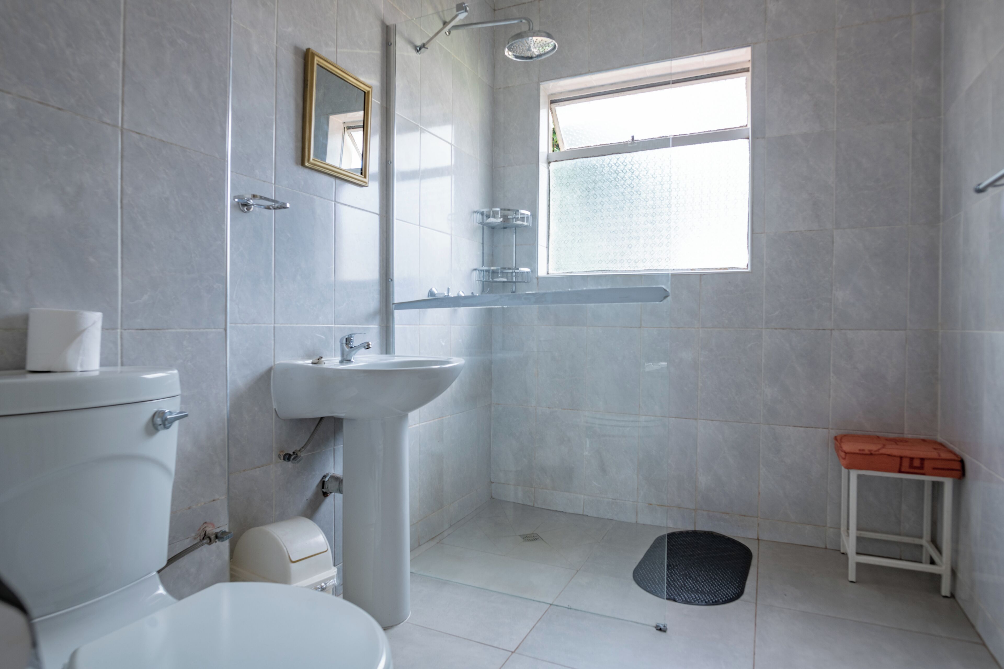 Baño con inodoro, lavamanos, ducha y un taburete pequeño.