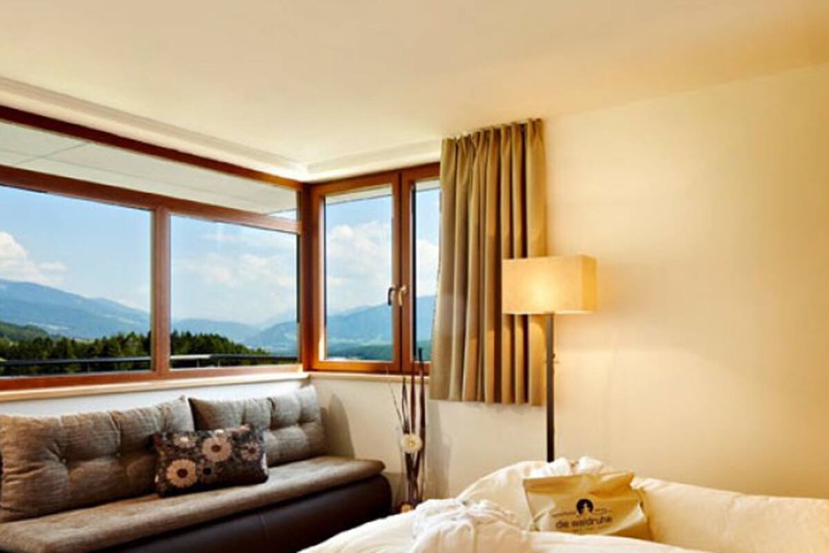 Habitación de hotel con cama, un sofá con cojines, una lámpara y vistas a montañas a través de grandes ventanales.