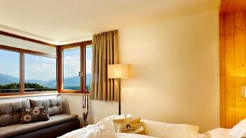 A hotel room with a bed, a sofa with cushions, a lamp, and a view of mountains through large windows.