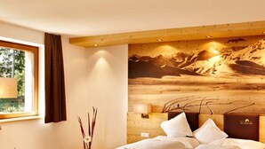Une chambre d’hôtel avec une tête de lit en bois, une grande fresque murale représentant un paysage de montagne, une fenêtre avec des rideaux et une table de chevet avec une lampe.