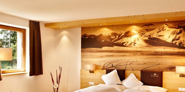 Une chambre d’hôtel avec une tête de lit en bois, une grande fresque murale représentant un paysage de montagne, une fenêtre avec des rideaux et une table de chevet avec une lampe.