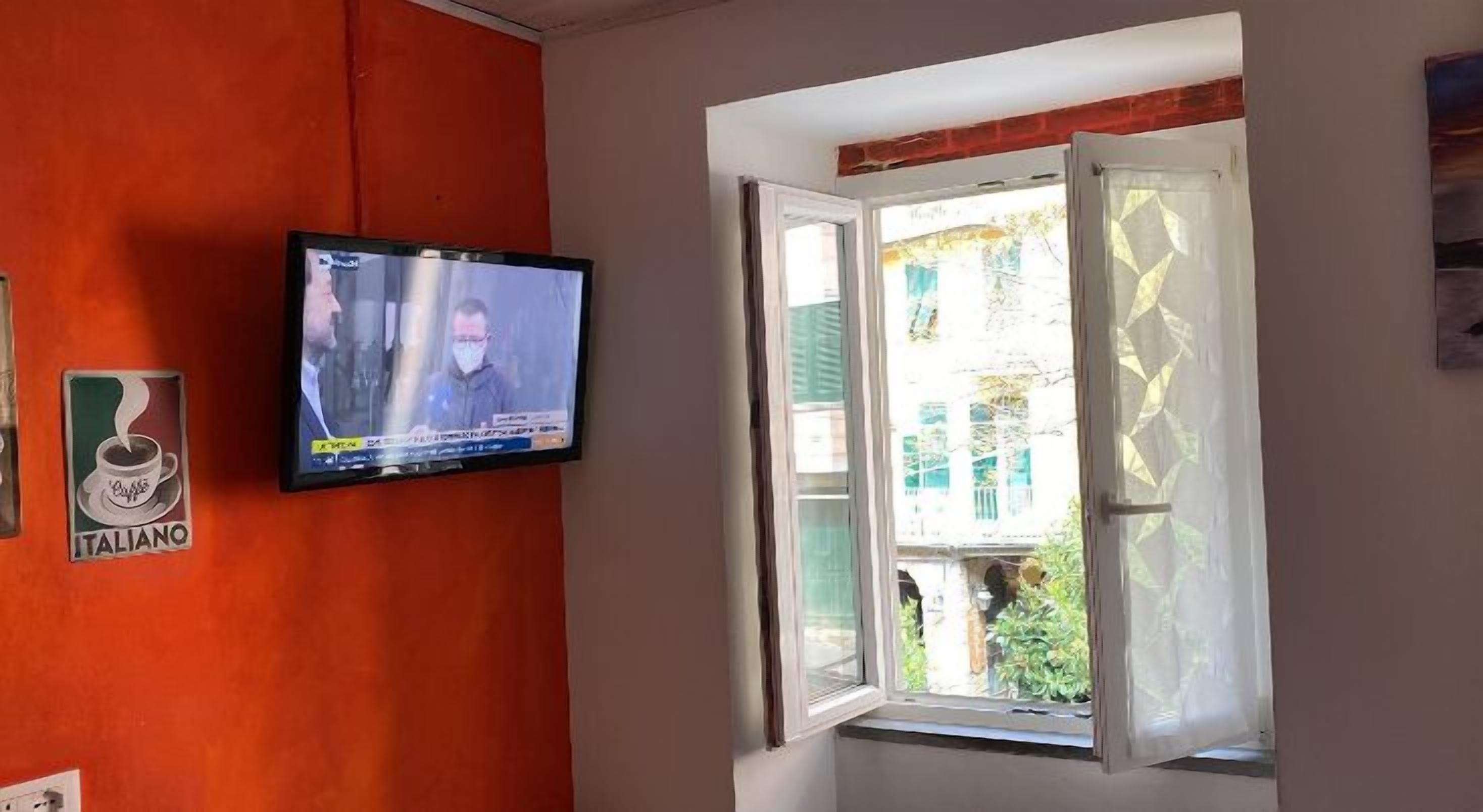 Una habitación con una televisión instalada en una pared naranja, una ventana con vistas a la vegetación exterior y un letrero que dice "ITALIANO".