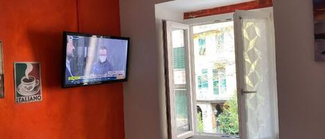 Una habitación con una televisión instalada en una pared naranja, una ventana con vistas a la vegetación exterior y un letrero que dice "ITALIANO".