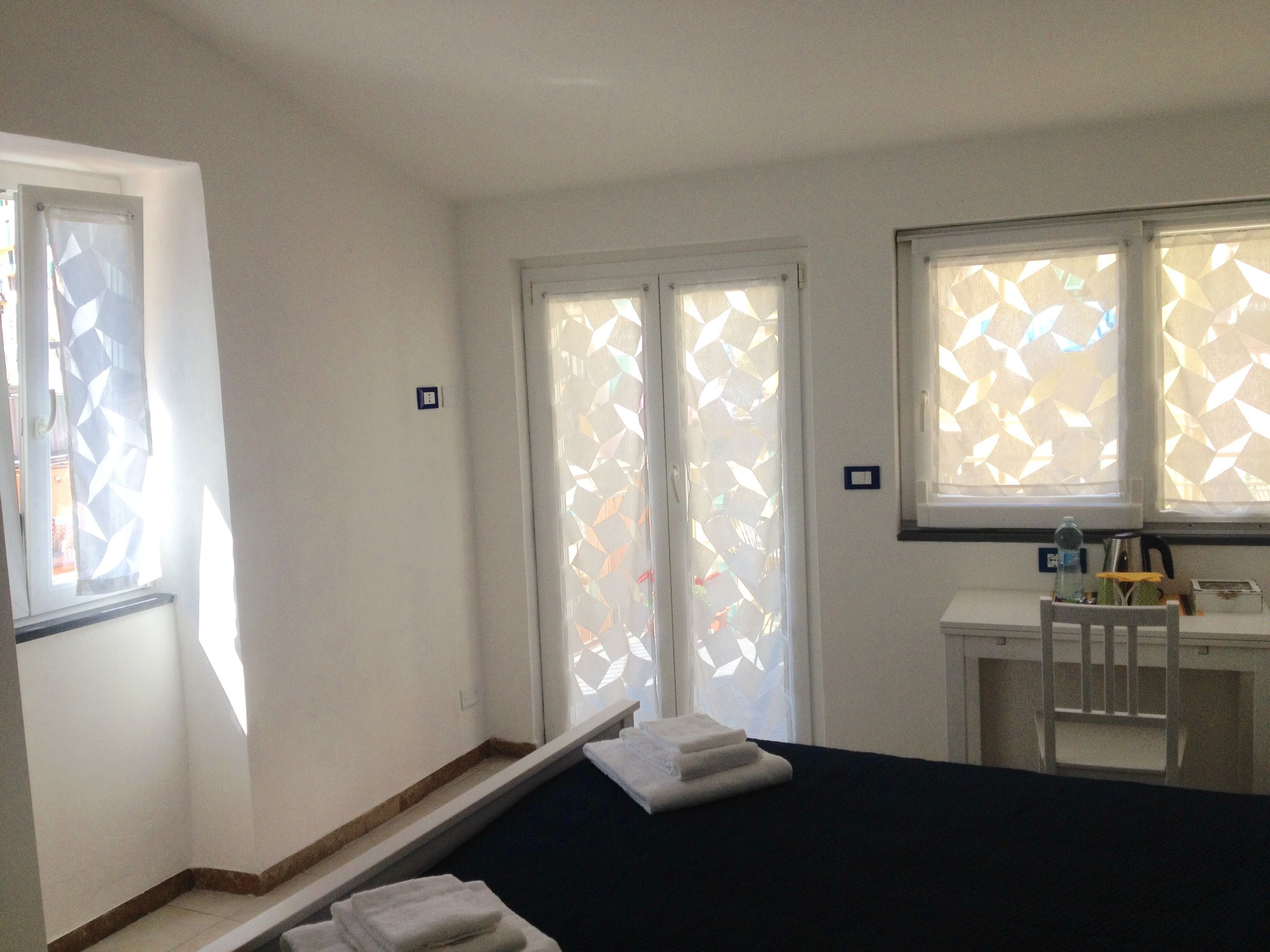 A hotel room with a bed, a desk, and a chair. There are two windows with geometric blinds.