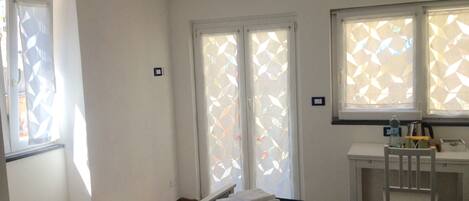 A hotel room with a bed, a desk, and a chair. There are two windows with geometric blinds.