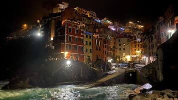 Vista notturna di una cittadina costiera con edifici illuminati, una costa rocciosa e un piccolo ponte sull'acqua.