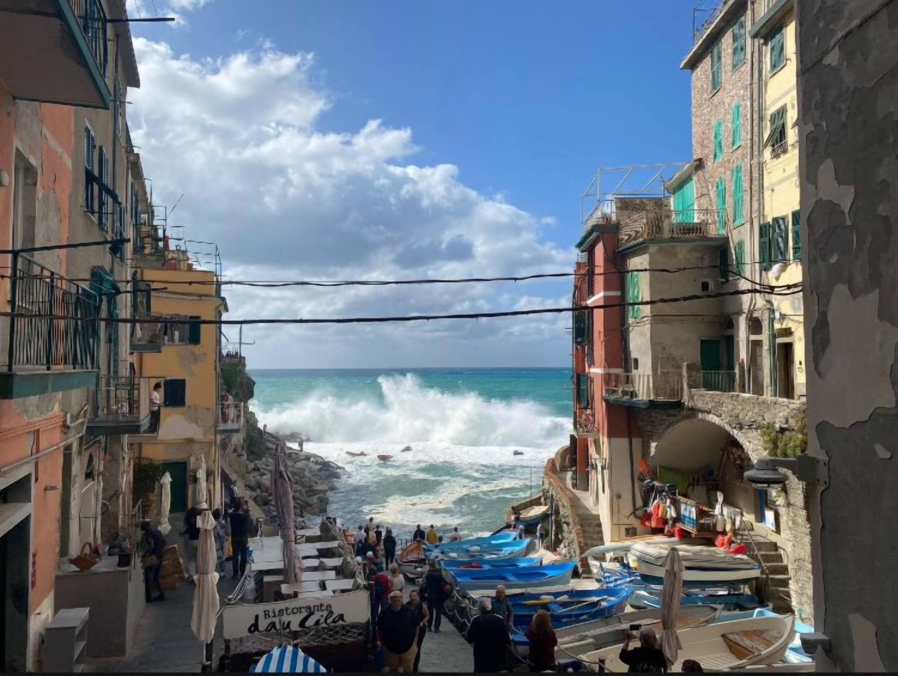 A coastal street with buildings on either side, a rocky shoreline, and several boats docked along the promenade.